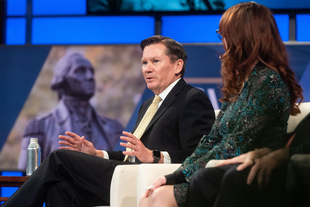 man speaking in chair wearing suit with george washington statue in background